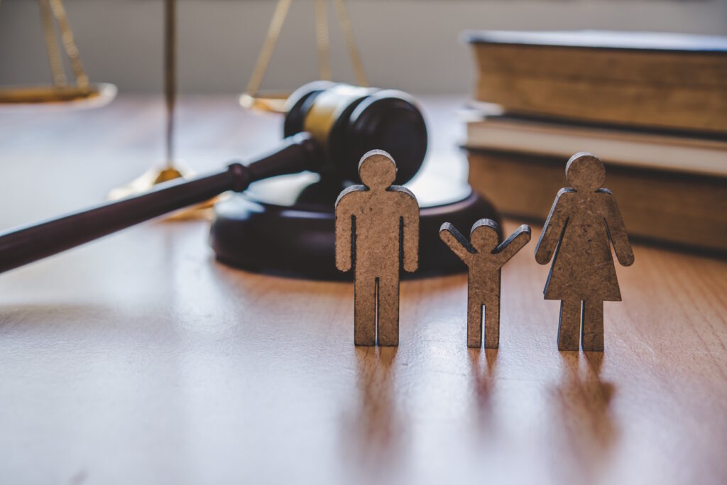 Wooden cutout figures of a family stand on a desk, with a blurred judge’s gavel and scales of justice in the background (legal/family law theme).