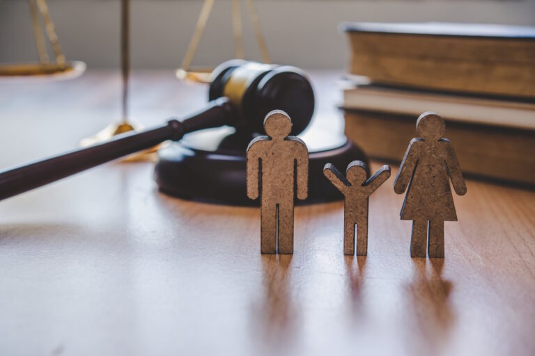 Wooden cutout figures of a family stand on a desk, with a blurred judge’s gavel and scales of justice in the background (legal/family law theme).