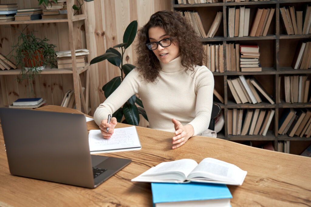 Woman with glasses writing in a notebook at a wooden desk, laptop nearby, in a study with tall bookshelves and a plant in the background.