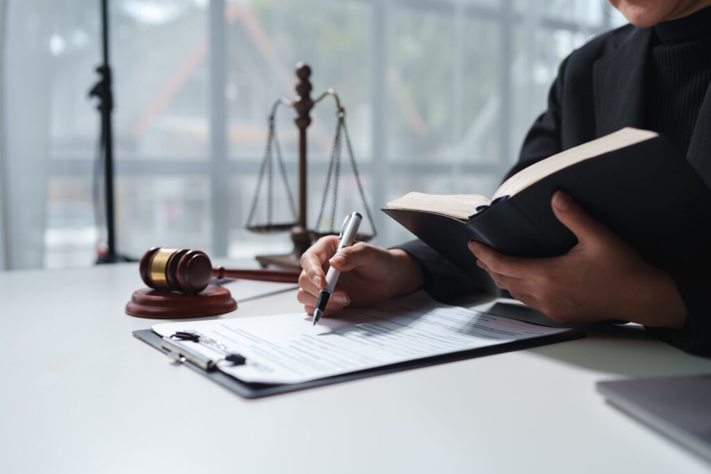 Gavel and scales of justice on a desk beside an open law book as a person in a suit signs documents.