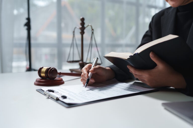 Gavel and scales of justice on a desk beside an open law book as a person in a suit signs documents.